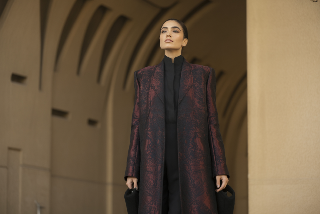 Editorial lifestyle fashion image of a middle eastern female model wearing a dramatic long structured coat inspired by opera aesthetics. Elegant posture, architectural background, cinematic lighting. Palette: black, deep wine, antique gold. High-end fashion editorial photography.