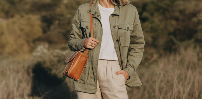 Lifestyle fashion photo of stylish white woman wearing khaki utility jacket with multiple pockets, white t-shirt underneath, beige linen trousers, tan leather crossbody bag, sunglasses, natural outdoor setting, golden hour lighting, safari chic aesthetic, professional lifestyle fashion photography, effortless adventure style