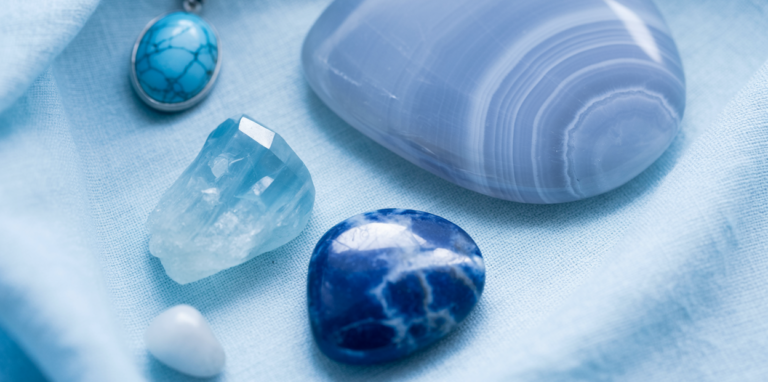 A close-up editorial flatlay of throat chakra crystals on a soft blue linen cloth — a pale blue lace agate palm stone, a small raw aquamarine piece, a sodalite tumbled stone, and a turquoise pendant arranged with generous space between them, soft natural light, bright and clear blue tones. Pinterest pin format cropped to top third. Photorealistic. No text. No words. No letters.
