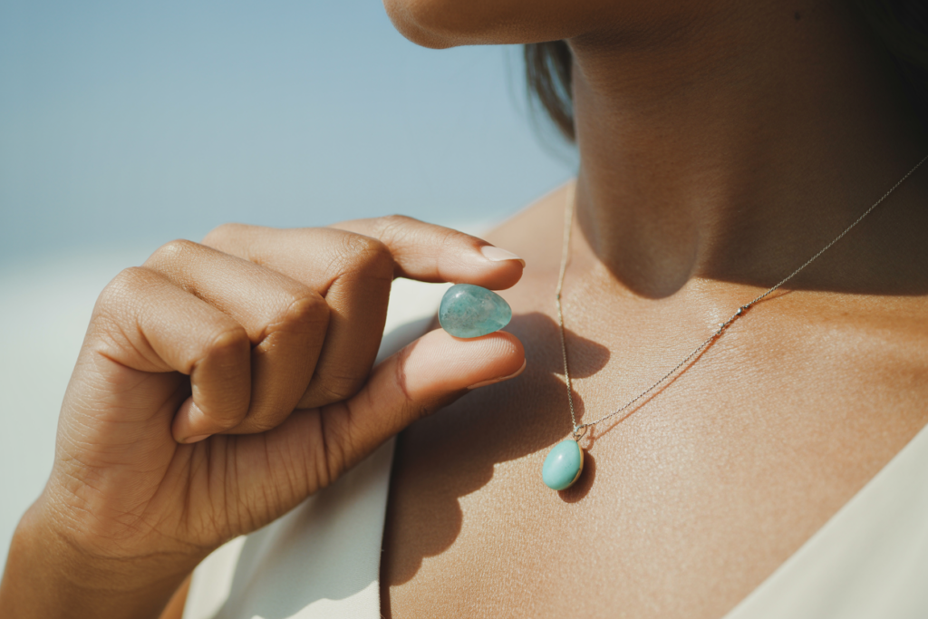 A close-up of a woman's hand with light brown skin holding a small aquamarine tumbled stone in a loosely closed fist, wearing a simple turquoise pendant necklace visible at the throat, soft blue and cream tones, warm natural light. Pinterest pin format cropped to bottom third. Photorealistic. No text. No words. No letters.
