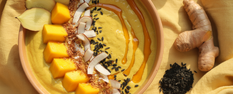 A stunning overhead editorial flatlay of a golden mango turmeric smoothie bowl in a wide shallow ceramic bowl in warm terracotta — deep golden yellow base with a swirl visible from blending, topped with fresh mango cubes arranged in a fan pattern, toasted coconut flakes scattered around, black sesame seeds sprinkled throughout, thin slices of fresh ginger at the edge, a drizzle of raw honey catching the light. Shot from directly above on a warm cream linen surface with a whole turmeric root and a small pile of black sesame seeds beside the bowl. Soft warm natural window light. Editorial luxury food photography aesthetic. Warm golden and cream tones. Pinterest pin format 2:3 vertical. Photorealistic. No text. No words. No letters.