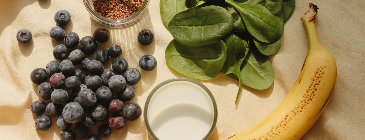 Editorial overhead flatlay of blueberry smoothie ingredients arranged on a warm cream linen surface — a pile of frozen wild blueberries, a handful of fresh spinach, a tablespoon of ground flaxseed, a small cup of almond milk, a halved banana. Soft natural light. Editorial luxury food photography aesthetic. Deep berry and cream tones. Pinterest pin format cropped to bottom third. Photorealistic. No text. No words. No letters.