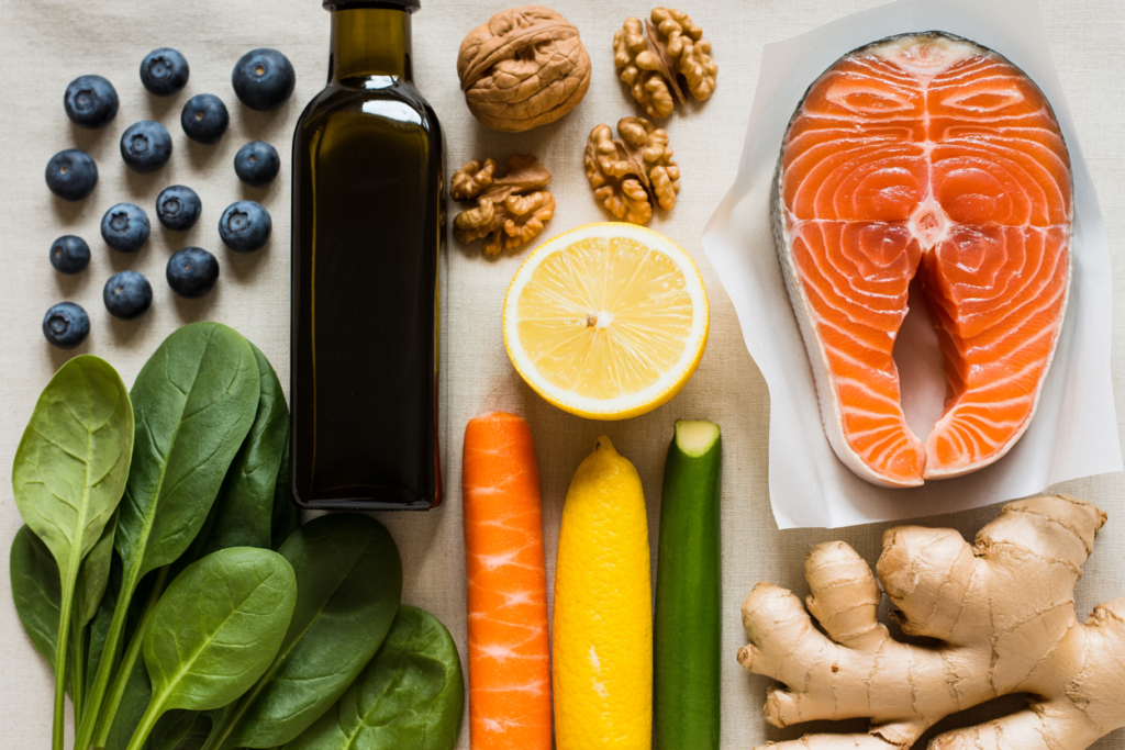 Editorial overhead shot of beautiful anti-inflammatory whole foods arranged on a warm cream linen surface — fresh blueberries, a dark bottle of extra-virgin olive oil, walnuts, a piece of fresh salmon, leafy spinach, colorful vegetables, a halved lemon, fresh ginger root. Bright warm natural lighting. Editorial luxury food photography aesthetic. Pinterest pin format cropped to bottom third. Photorealistic. No text. No words. No letters.