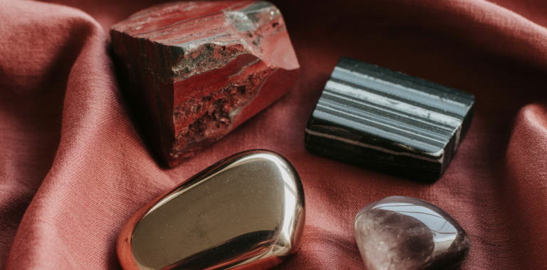 Editorial flatlay of root chakra crystals on a deep burgundy linen cloth — a raw red jasper chunk, a striated black tourmaline piece, a smooth metallic hematite palm stone, and a smoky quartz tumbled stone arranged with generous editorial space between them. Warm earth tones and deep red natural light. Pinterest pin format cropped to top third. Photorealistic. No text. No words. No letters.