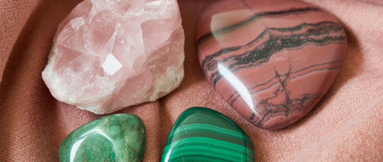 Editorial flatlay of heart chakra crystals on a soft dusty rose linen cloth — a large rose quartz raw chunk, a rhodonite palm stone showing its distinctive pink and black pattern, a green aventurine tumbled stone, and a small malachite piece arranged with generous editorial space between them. Soft warm natural light. Rose and green tones. Pinterest pin format cropped to top third. Photorealistic. No text. No words. No letters.
