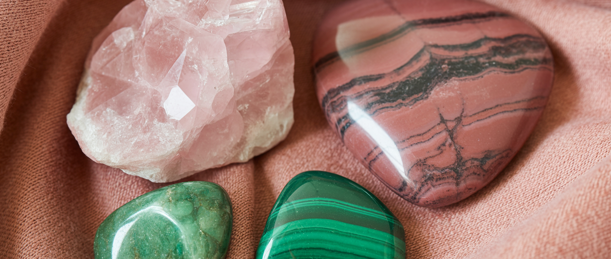 Editorial flatlay of heart chakra crystals on a soft dusty rose linen cloth — a large rose quartz raw chunk, a rhodonite palm stone showing its distinctive pink and black pattern, a green aventurine tumbled stone, and a small malachite piece arranged with generous editorial space between them. Soft warm natural light. Rose and green tones. Pinterest pin format cropped to top third. Photorealistic. No text. No words. No letters.