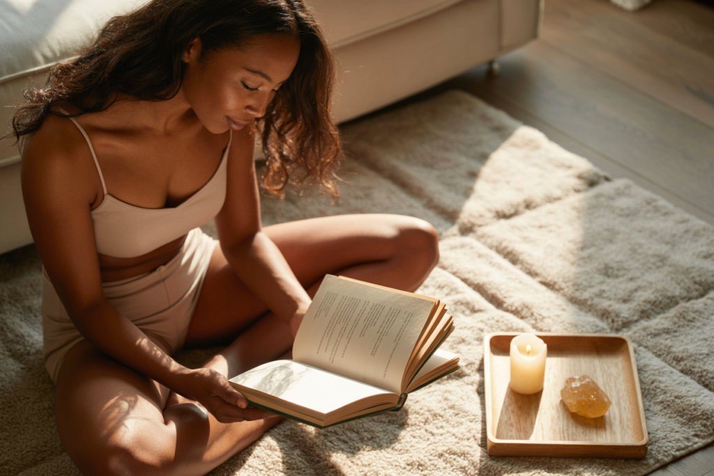 A light-skinned Black woman in her 30s with long hair, sitting cross-legged on a cream rug in soft warm morning light, holding a beautiful chakra healing book open in her lap, looking down at it with quiet curiosity and openness — not performing spirituality, just genuinely reading and considering. A small citrine crystal and a lit beeswax candle on a low tray beside her. Warm cream and golden tones. Cozy editorial luxury wellness lifestyle aesthetic. Full body shot head to toe. Pinterest pin format 2:3 vertical. Photorealistic. No text. No words. No letters.