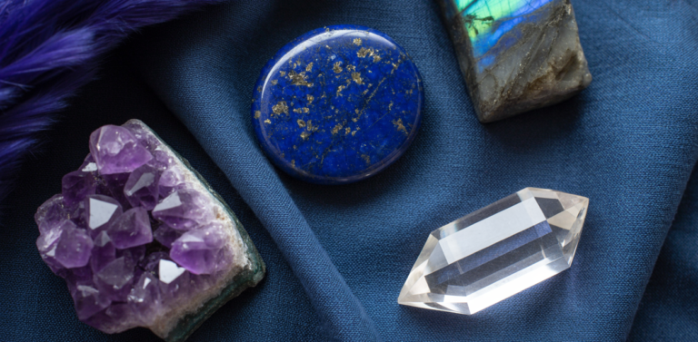 Editorial flatlay of third eye chakra crystals on a deep indigo linen cloth — a raw amethyst cluster, a smooth lapis lazuli palm stone showing deep midnight blue with gold pyrite flecks, a labradorite freeform piece catching iridescent light, and a clear quartz point arranged with generous editorial space between them. Deep indigo and purple natural light. Pinterest pin format cropped to top third. Photorealistic. No text. No words. No letters.