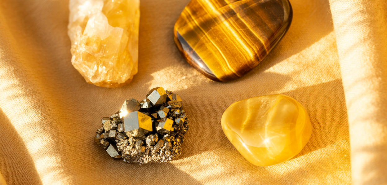 Editorial flatlay of solar plexus crystals on a warm golden linen cloth — a natural citrine raw point, a tiger's eye palm stone showing its distinctive gold and brown striations, a small pyrite cluster catching the light, and a yellow jasper tumbled stone arranged with generous editorial space between them. Warm golden natural light. Pinterest pin format cropped to top third. Photorealistic. No text. No words. No letters.
