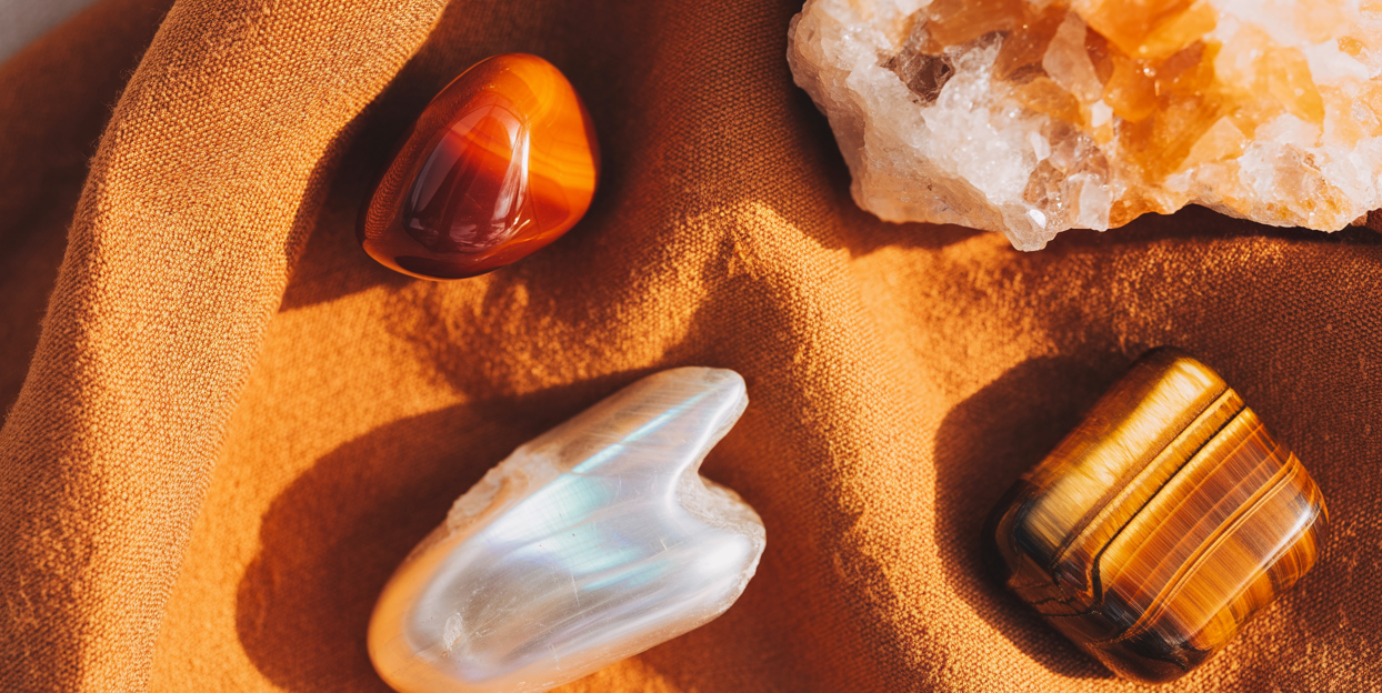 Editorial flatlay of sacral chakra crystals on a warm terracotta linen cloth — a deep orange carnelian tumbled stone, a luminous pale orange calcite raw chunk, a pearlescent moonstone palm stone, and a tiger's eye tumbled piece arranged with generous editorial space between them. Warm amber natural light. Pinterest pin format cropped to top third. Photorealistic. No text. No words. No letters.