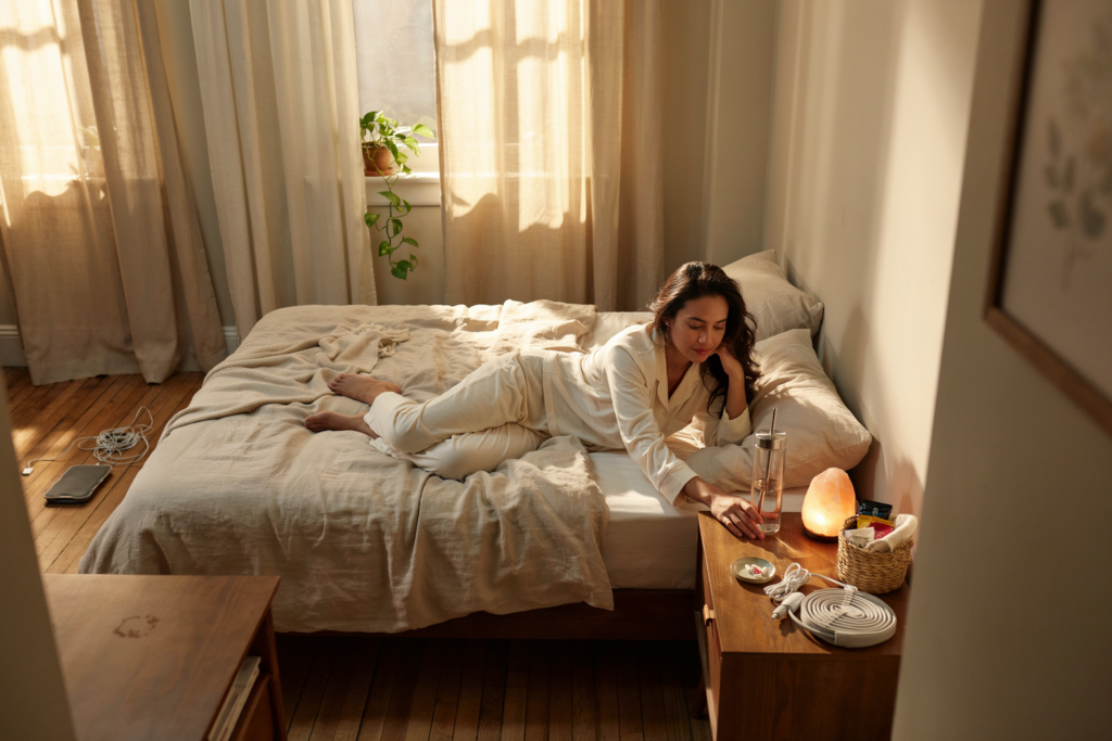 Wide shot from across the room at bed height. Two visual zones visible in the same frame — left side of the bed: nightstand bare, heating pad on the floor out of reach, phone face-down across the room. Right side: nightstand fully set up — water bottle with straw, snack basket, salt lamp, heating pad coiled and ready. Latino woman visible on the right side, hand resting on the nightstand naturally. Warm late morning light from a window behind, golden and low. Emotional quality: the concrete difference preparation makes. Shot on 35mm, editorial lifestyle, warm and documentary. No text in the image.