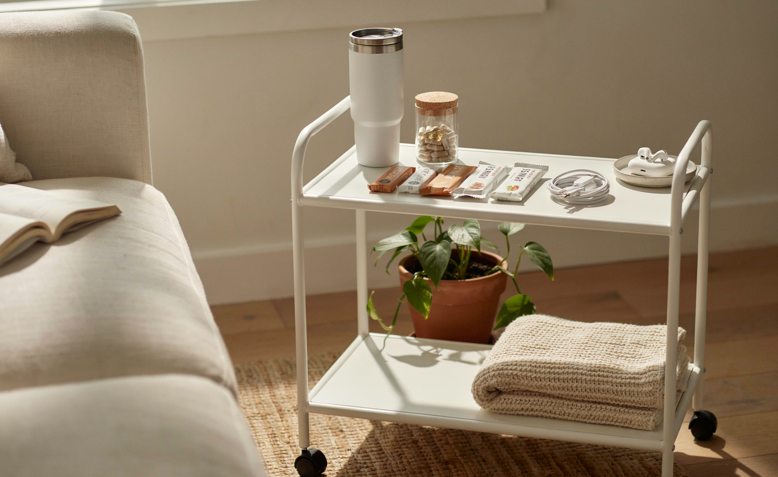 A close-up editorial shot of a white three-tier rolling cart positioned beside a cream sofa — top tier holding a large insulated tumbler and a small jar of supplements, middle tier with individually wrapped snacks and a phone charger coiled neatly, bottom tier with a soft folded throw blanket. Everything organized and within arm's reach of someone lying on the sofa. Warm cream and white tones. Soft natural light. Clean and intentional. Pinterest pin format 2:3 vertical. Photorealistic. No text. No words. No letters.
