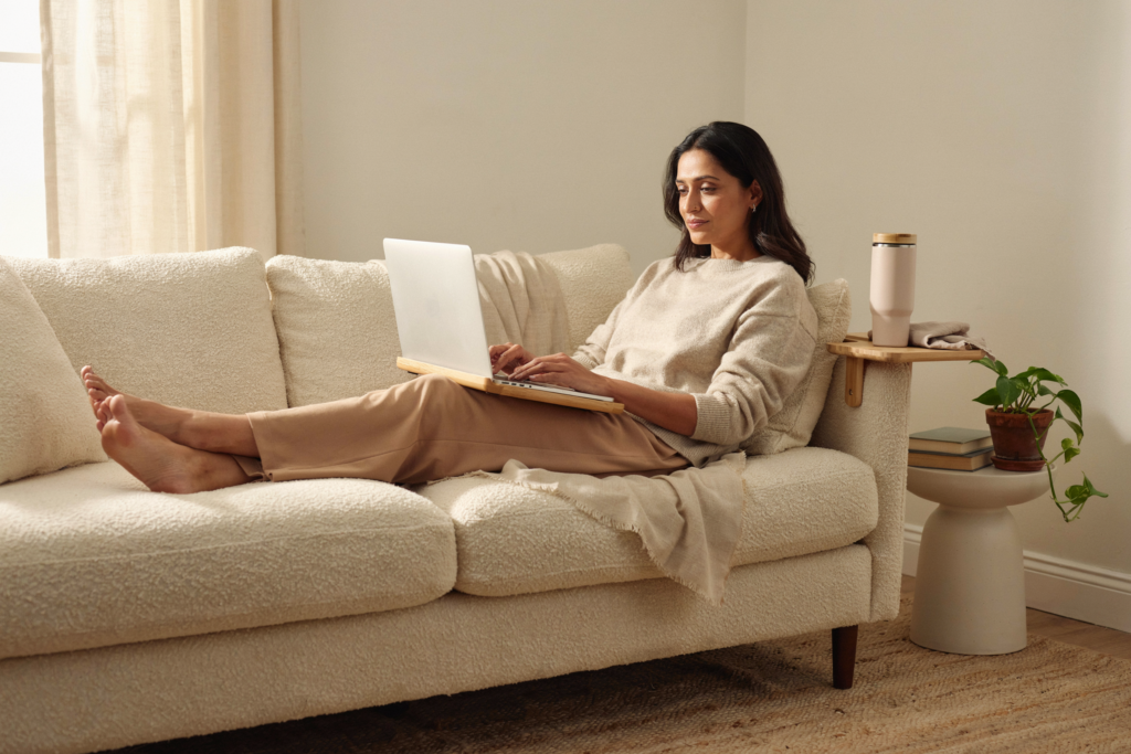 A South Asian woman in her early 40s lying on a sofa with a lap desk across her thighs, a laptop open on the desk, a large insulated tumbler within reach on a clip-on sofa arm tray beside her, expression calm and settled — everything she needs is right there, no getting up required. She looks like someone who has figured something out. Warm cream and soft neutral tones. Natural afternoon light. Editorial luxury wellness lifestyle aesthetic. Full body shot head to toe. Pinterest pin format 2:3 vertical. Photorealistic. No text. No words. No letters.