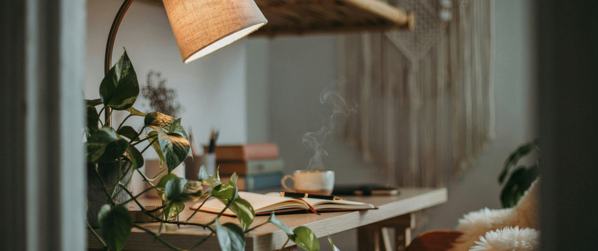 Low angle shot from below desk height, camera angled slightly upward. A boho home office scene from an intimate vantage point — bamboo desk surface above, warm lamp light spilling down from the left, trailing pothos cascading over the desk edge toward the camera, sheepskin throw visible on the chair arm. The angle makes the workspace feel enclosing and warm — like being held inside the room rather than observed from outside it. No person in frame. Emotional quality: shelter. Shot on 35mm, cinematic warm tones, depth of field with sharp foreground plant and softly glowing background. No text in the image. No text in the image.