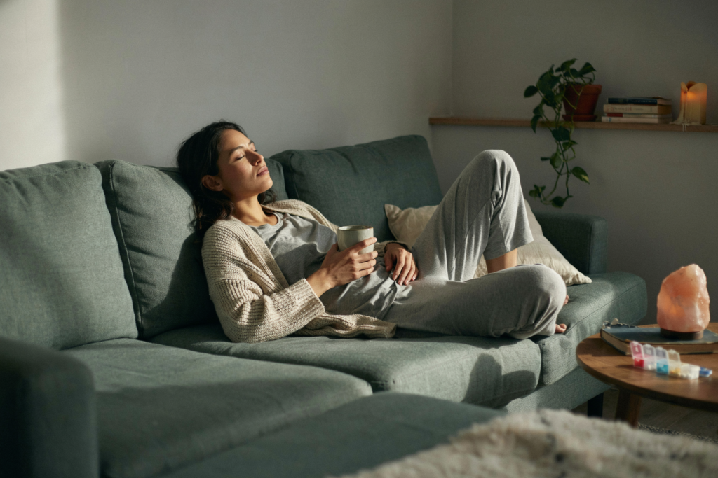 Mid-shot from slightly above, camera angled gently downward. A woman lying on a sofa under a chunky knit throw, Sunday afternoon light falling across her from a window to the left. One hand holds a mug resting on her chest, the other is relaxed at her side. Eyes closed, expression peaceful rather than exhausted — resting with permission rather than collapse. A small side table beside the sofa: salt lamp glowing, a book face-down, a pill organizer. Latino woman with dark hair loose across the cushion. Soft diffused window light, warm and low. Emotional quality: rest as a radical act of preparation. Shot on medium format film, warm editorial, documentary intimacy. No text in the image.