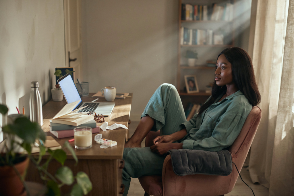 Cinematic wide shot, 30-degree angle from slightly elevated. A Black woman with long hair— deep brown skin, wearing a soft sage linen set — seated at a home desk, laptop open, but not working. She is looking away from the screen, toward the window beside her. The desk around her is lived-in — a water bottle, a heating pad draped over the chair arm, a pill organizer on the corner. Outside the window, afternoon light. Physical drama: the laptop screen casts a cool blue-white glow across the desk surface while warm afternoon light from the window falls across her face and shoulder simultaneously — two competing light qualities, the work and the rest, both present, neither fully winning. Emotional specificity: her gaze out the window carries the very specific quality of someone recalibrating — not avoiding the work, not resting, but sitting in the gap between what working from home was supposed to be and what it actually is. No text anywhere in the image. Shot on medium format film, cinematic, warm afternoon and cool screen light.