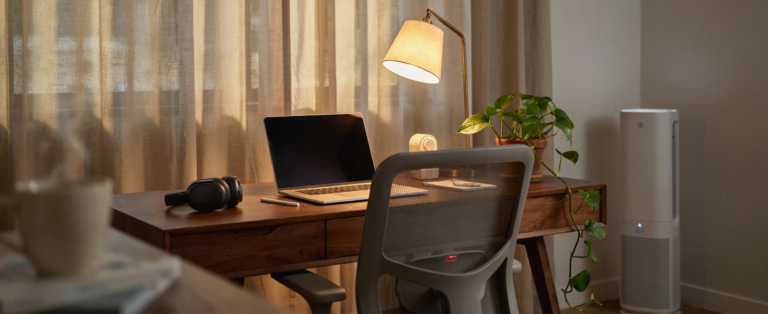 wfh desk setup - Warm amber desk lamp at eye level replacing the overhead fluorescent. A white noise machine on the corner of the desk. ergonomatical chair , with heat pad , noise cacelling headphones Linen curtains filtering the afternoon sun into something golden rather than glaring. A small pothos plant on the desk corner catching the warm lamp light. An air purifier running quietly beside the desk. the warm desk lamp creates a contained pool of amber light across the desk surface — the screen, the keyboard, all in the same warm light, the rest of the room softly lit rather than harshly illuminated. The linen curtain filters the window light into something warm and diffuse. No text anywhere in the image. Shot on medium format film, warm amber desk lamp.