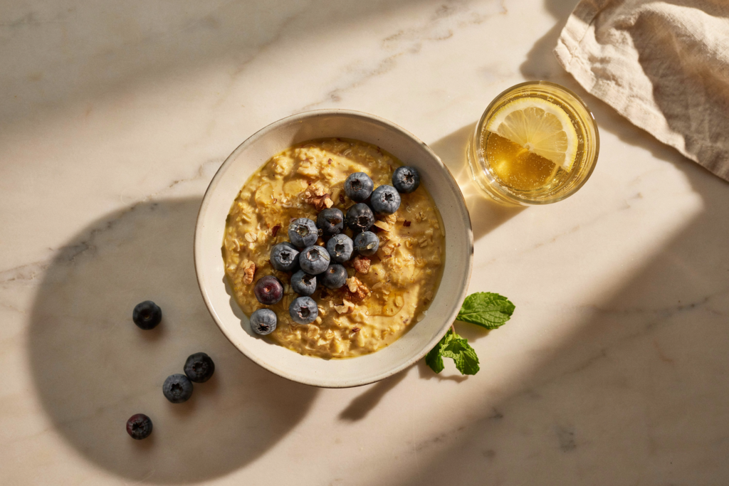 Overhead flat lay, camera directly above. A styled breakfast scene on a warm cream marble surface — a wide ceramic bowl of golden turmeric overnight oats topped with fresh blueberries, a drizzle of honey, and crushed walnuts. Beside it: a small glass of golden turmeric water with a lemon slice, a few loose blueberries scattered naturally, a sprig of fresh mint. Warm morning side light from the left — golden, directional, one side in soft shadow. Editorial luxury food photography. Rich color contrast between the golden oats and deep purple blueberries. No hands. No person. No mug. Photorealistic. No text.