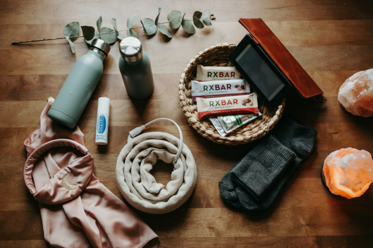 Overhead flat lay shot directly above a styled nightstand surface. Everything arranged with intentional ease: insulated water bottle with straw, RXBAR and electrolyte packets in a small woven basket, cordless heating pad coiled neatly, Kindle e-reader, sleep headband, small tube of Biofreeze roll-on, fuzzy socks folded beside the basket, salt lamp glowing at the frame's edge. Warm side lighting from just outside the frame — golden and directional. No person in frame. Objects arranged as though someone prepared this space for themselves with care. Emotional quality: the quiet power of being ready. Shot on 35mm overhead, editorial still life, warm and intimate. No text in the image.