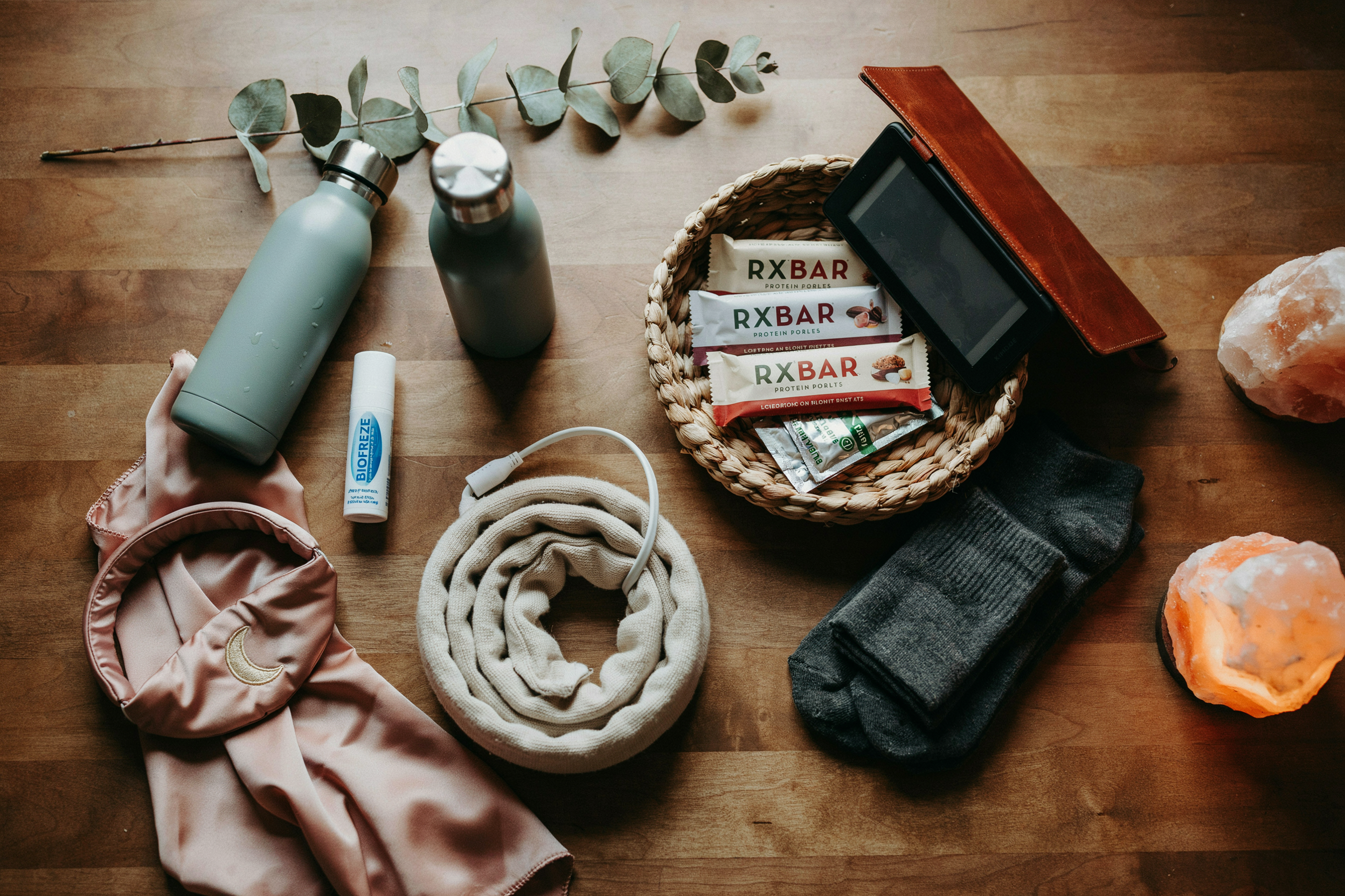 Overhead flat lay shot directly above a styled nightstand surface. Everything arranged with intentional ease: insulated water bottle with straw, RXBAR and electrolyte packets in a small woven basket, cordless heating pad coiled neatly, Kindle e-reader, sleep headband, small tube of Biofreeze roll-on, fuzzy socks folded beside the basket, salt lamp glowing at the frame's edge. Warm side lighting from just outside the frame — golden and directional. No person in frame. Objects arranged as though someone prepared this space for themselves with care. Emotional quality: the quiet power of being ready. Shot on 35mm overhead, editorial still life, warm and intimate. No text in the image.