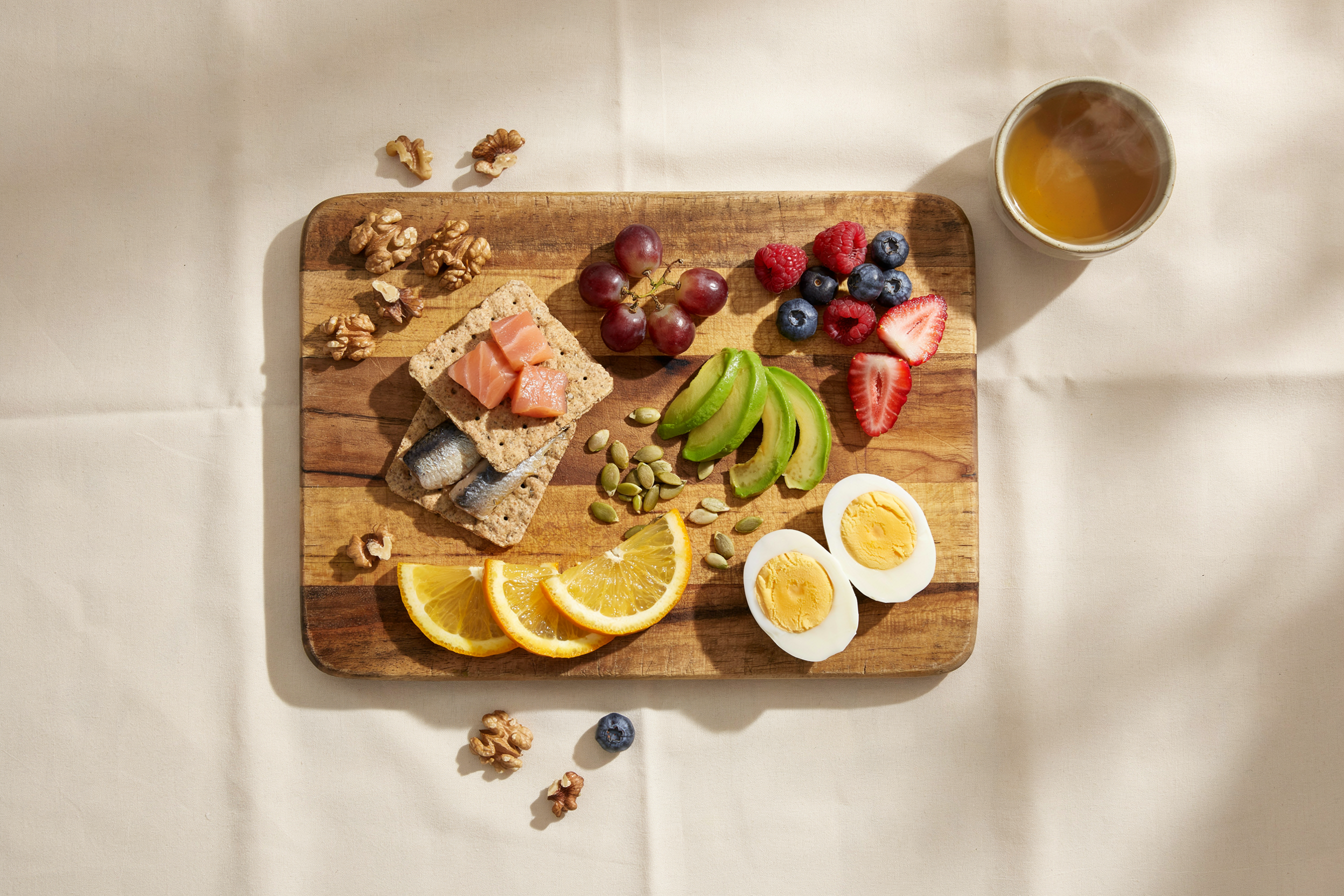 Overhead flat lay directly above. A low-effort anti-inflammatory BREAKFAST on a small wooden cutting board — a handful of walnuts, a small cluster of dark grapes, berries, orange slices , two brown rice crackers, a few cubes of salmon or sardine on crackers, a small avocado slices, teas, a sprinkle of pumpkin seeds, a boiled egg. Board sits on a warm cream linen surface. Warm natural side light, golden and soft. Editorial food photography — abundant but unfussy, the kind of board assembled from what's already in the kitchen. No hands. No person. Photorealistic. No text.