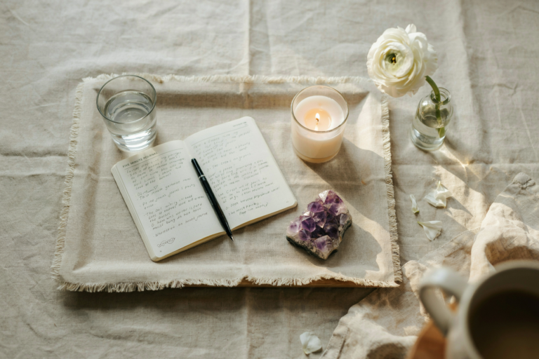 Overhead flat lay shot directly above a styled Sunday reset surface. A linen tray holding: a small glass of water, an open Five Minute Journal with a pen resting across it, a lit soy candle, a small amethyst crystal, a single fresh flower in a bud vase. Warm side lighting from a window just outside the frame — golden and soft. No hands, no person. Objects arranged with unhurried ease, nothing rigidly symmetrical. Emotional quality: intention without effort. Shot on 35mm overhead, editorial still life, warm and intimate. No text in the image.