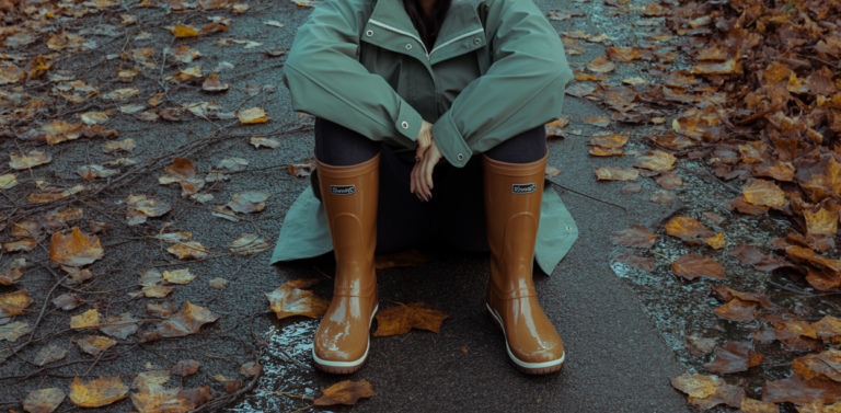 Editorial fashion photograph of a woman in waterproof duck boots styled with a rain jacket, standing on a wet leaf-covered path, moody overcast autumn light, no text