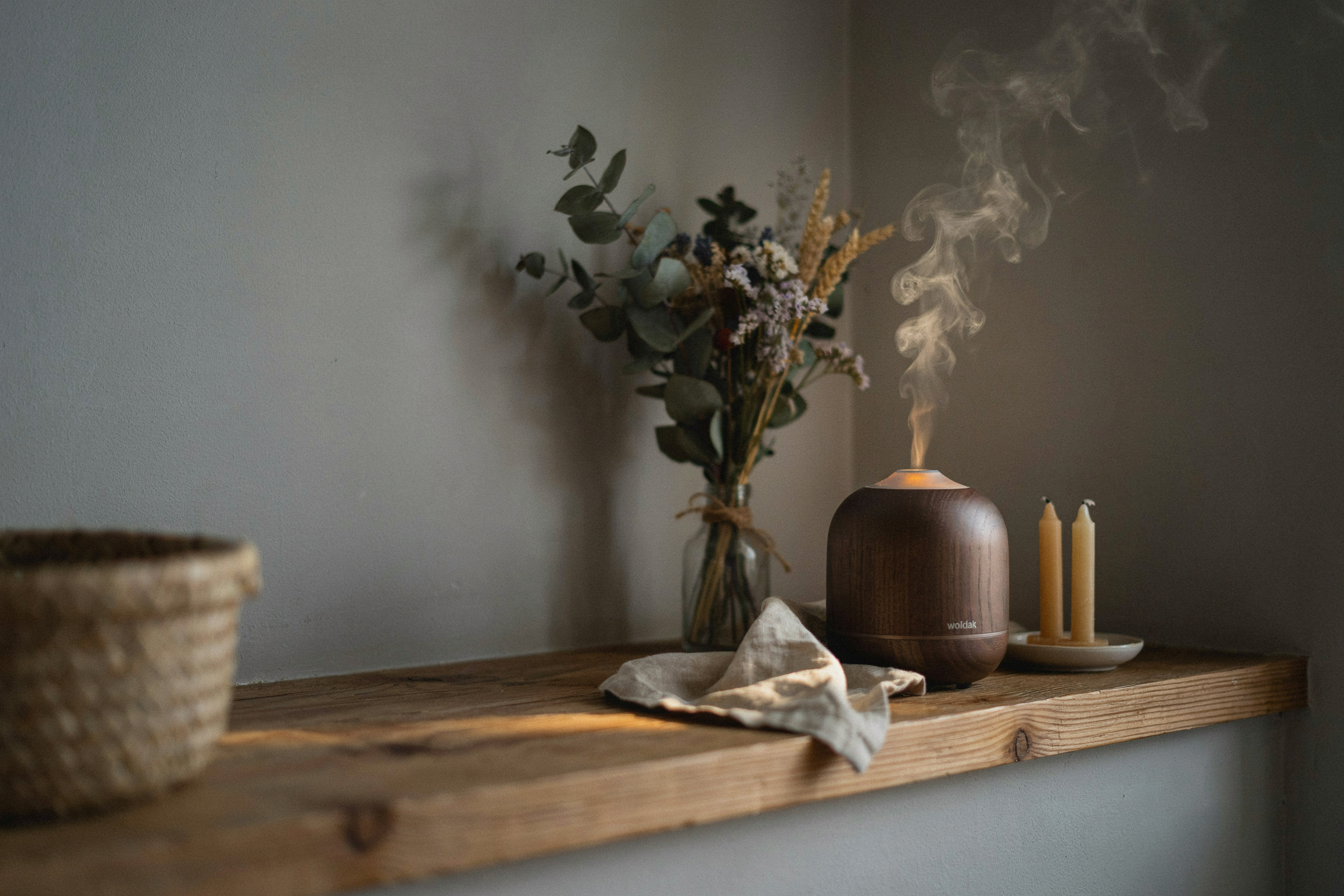 Editorial close-up photograph of a wood-finish essential oil diffuser releasing visible mist on a wooden shelf, warm ambient evening light, dried botanical stems softly blurred in background, no people, no text