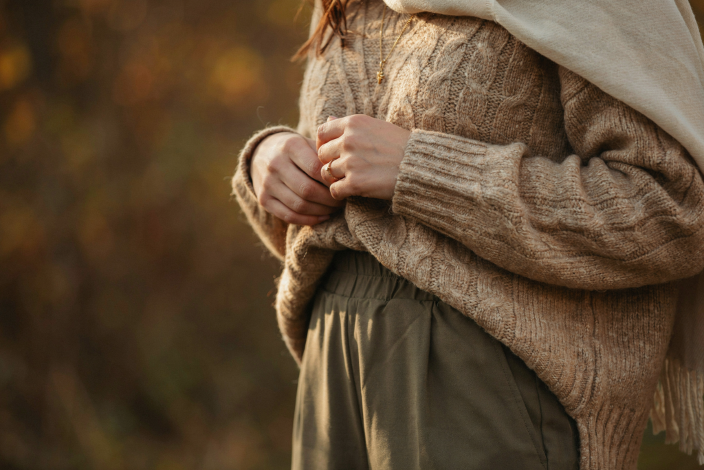 An extremely close editorial detail shot — a woman's midsection and hands, warm brown skin, wearing a chunky oversized camel knit sweater tucked loosely at one side into wide-leg olive trousers with a barely-visible elastic waistband, one hand in a deep pocket, a delicate gold chain necklace visible at the collarbone, a soft blanket scarf beginning at the shoulder. The image is cropped tightly — no face, no feet — purely the texture and detail of the outfit. Warm golden fall light. High fashion editorial aesthetic. Pinterest pin format 2:3 vertical. Photorealistic. No text. No words. No letters.