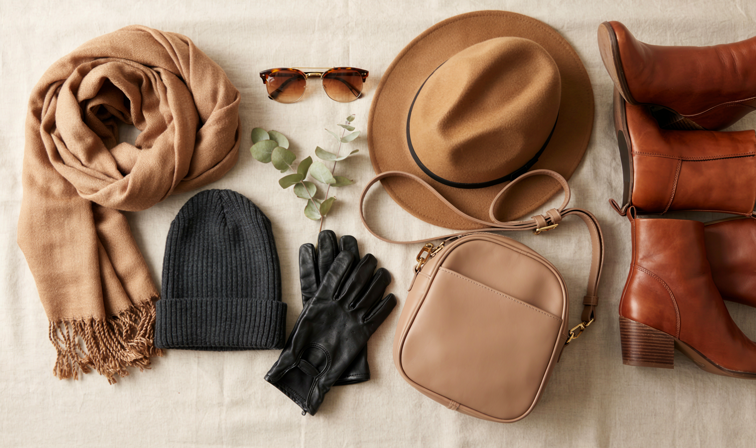 Fashion accessories arranged :, scarf, fadora hat, beanie , fingerless gloves, boots, sunglasses, a vegan leather crossbody bag with a wide padded strap ,minimalist flat lay.