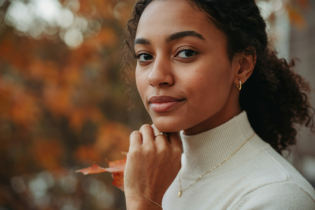a black woman wearing minimal danity gold jewelry up close necklace, earrings, bracelet, and ring , fall season