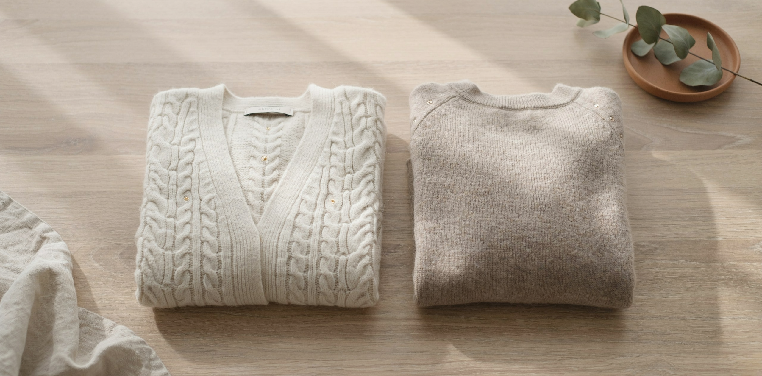 Two cocoon cardigans folded neatly on a wooden table, bright natural light filling the room, soft shadows, cozy and minimal style, realistic yarn texture, top-down view, warm and airy atmosphere, no text